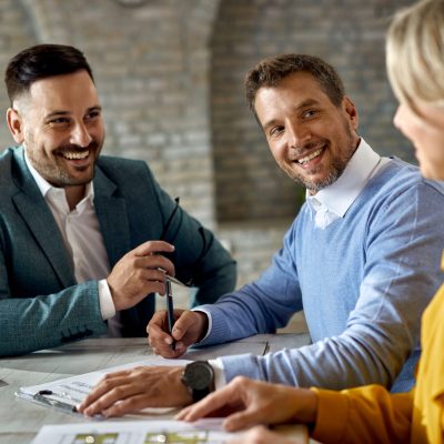 Happy man signing a contract while being with his wife on a meet Happy man talking to his wife while signing an agreement with insurance agent in the office.